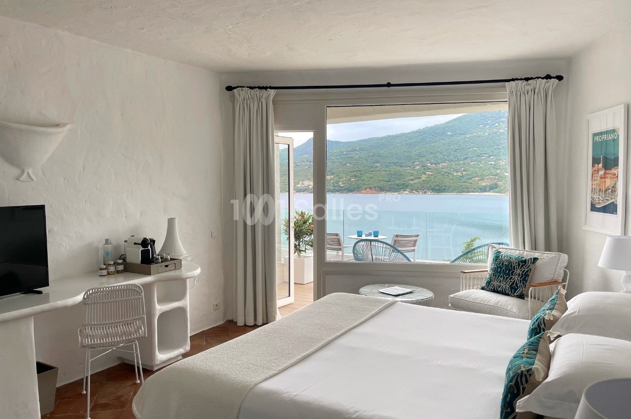 Chambre lumineuse avec lit double, bureau, fauteuil et vue sur une terrasse donnant sur la mer et les collines.