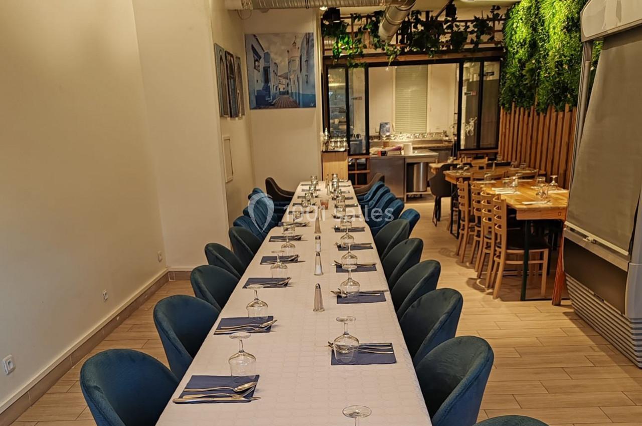 Salle de restaurant avec une longue table dressée, chaises bleues, décoration végétale et lumière naturelle.