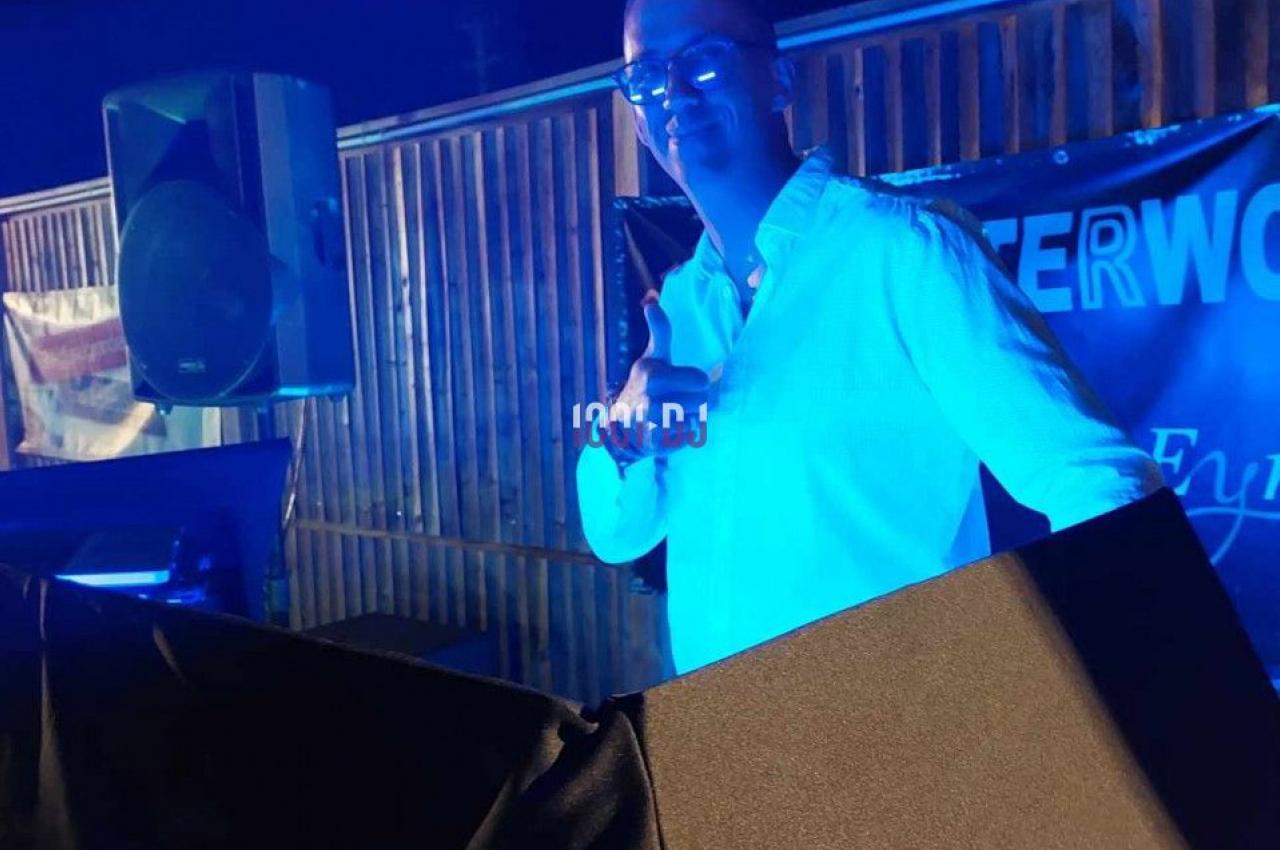 Un homme en chemise blanche se tient derrière une table de mixage sous un éclairage bleu, en extérieur.