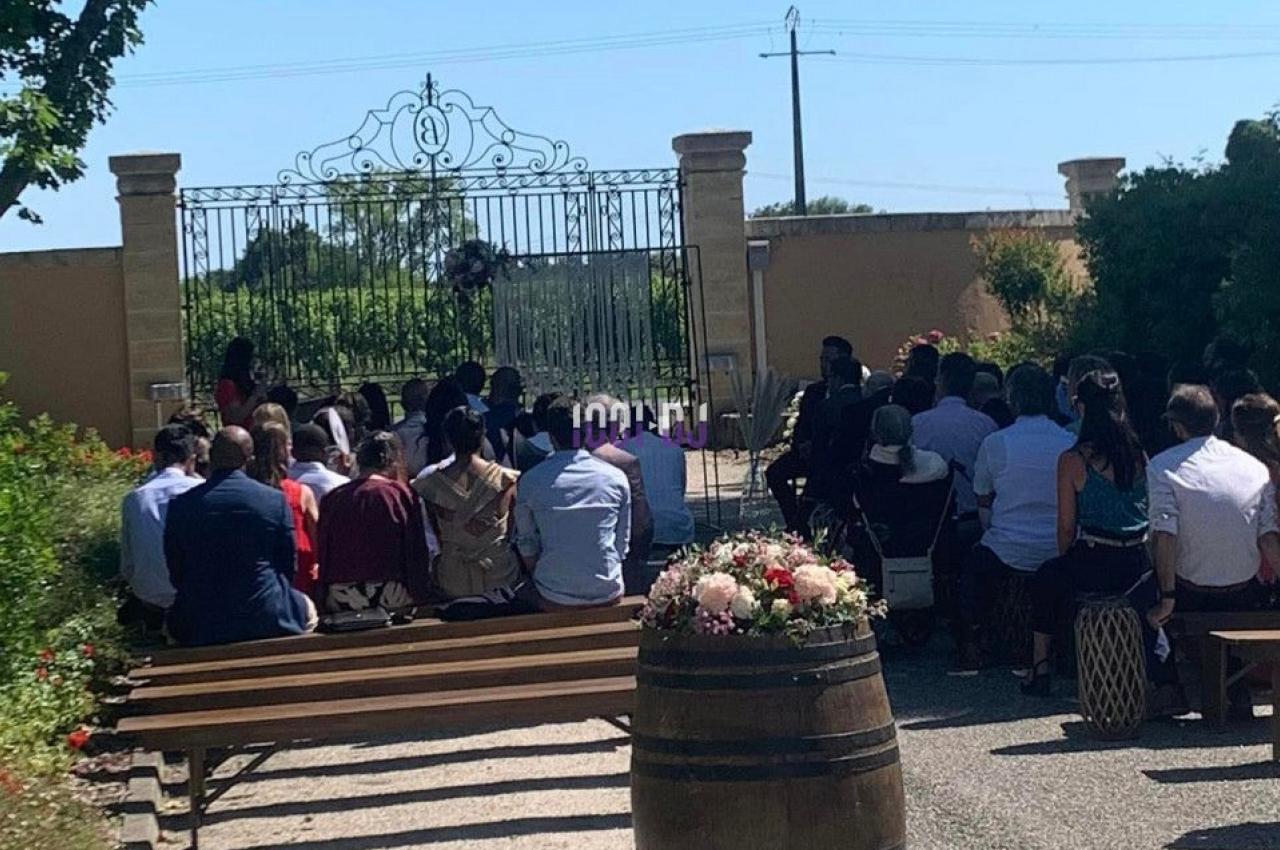 Des personnes assistent à une cérémonie en plein air, assises sur des bancs face à un portail en fer forgé.