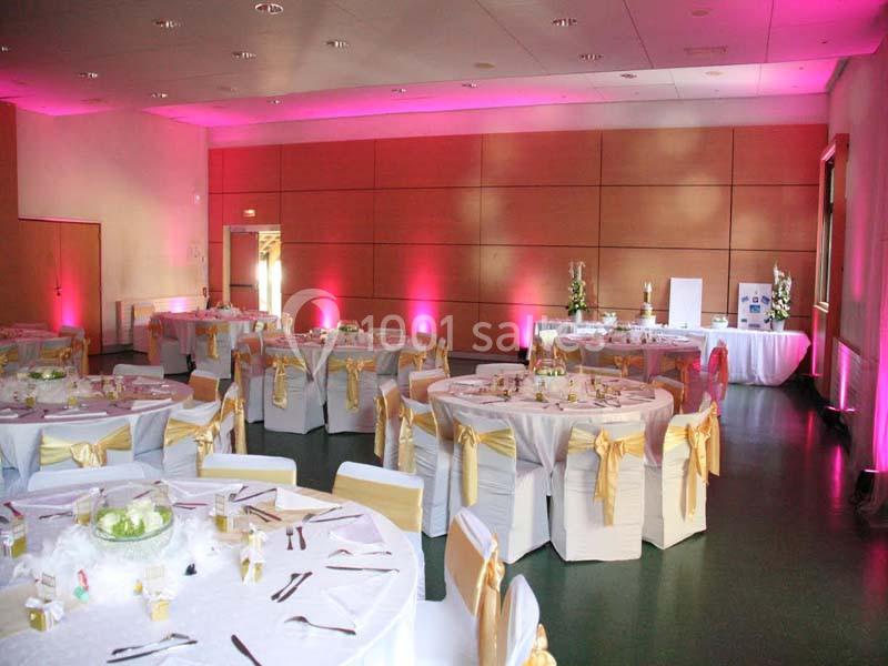 Salle de réception décorée avec des nappes blanches, des chaises ornées de rubans dorés et un éclairage rose.