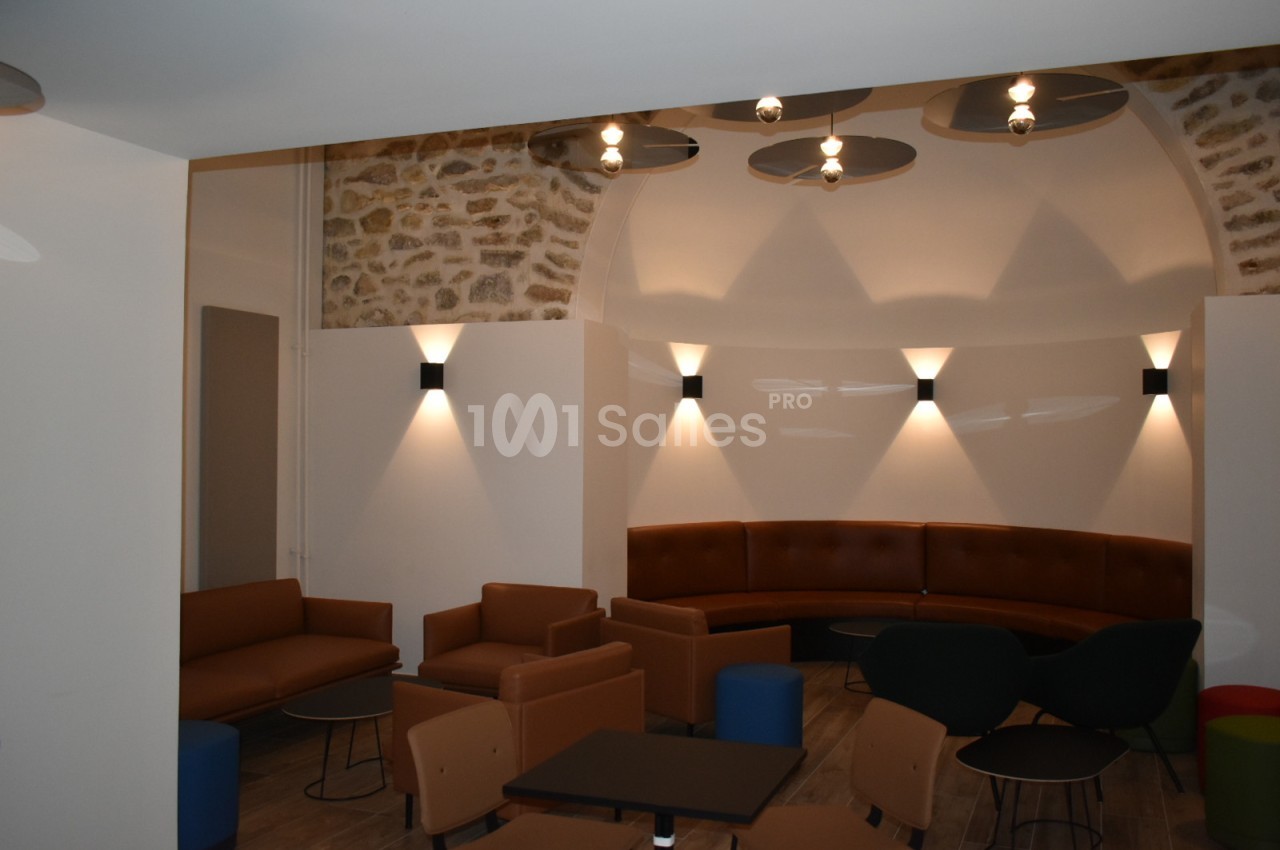 Espace intérieur avec fauteuils, tables basses et éclairage mural dans une pièce aux murs en pierre et plafond voûté.