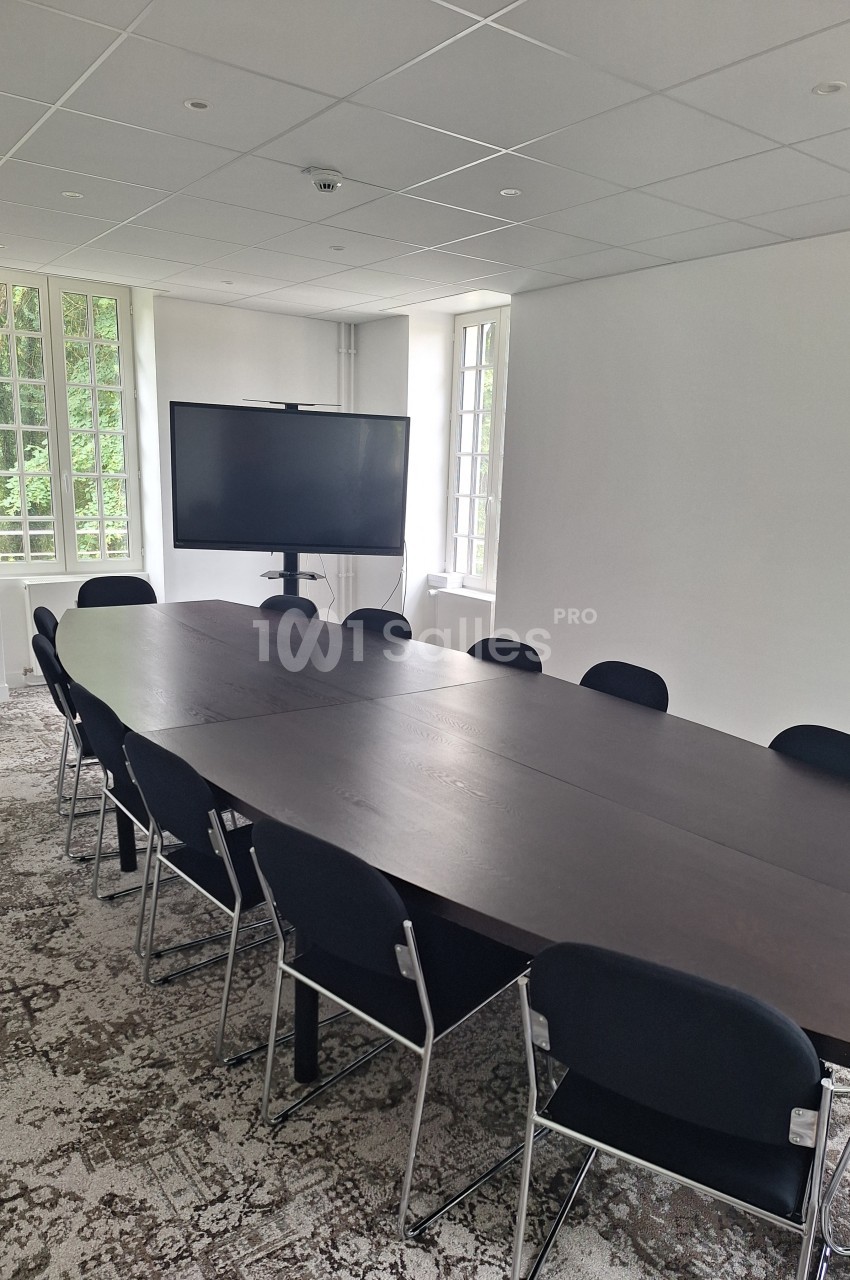 Salle de réunion lumineuse avec une grande table en bois foncé, des chaises noires et un écran sur pied.