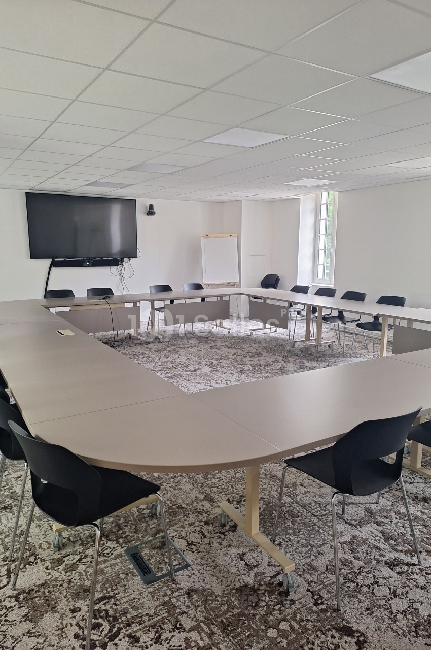 Salle de réunion lumineuse avec tables disposées en U, chaises noires, écran mural et tableau blanc.