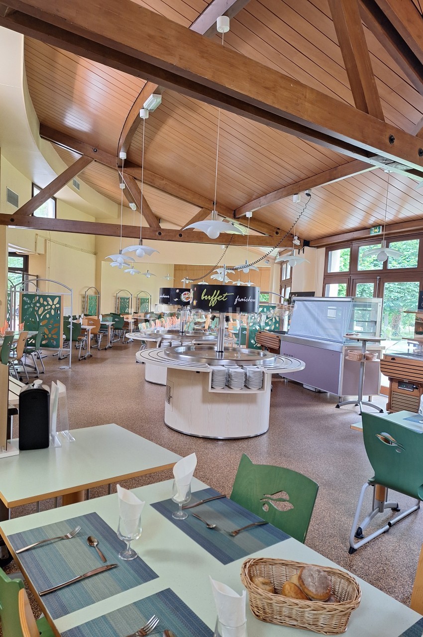 Salle de restaurant lumineuse avec tables dressées, buffet central et mobilier en bois sous un plafond en poutres apparentes.