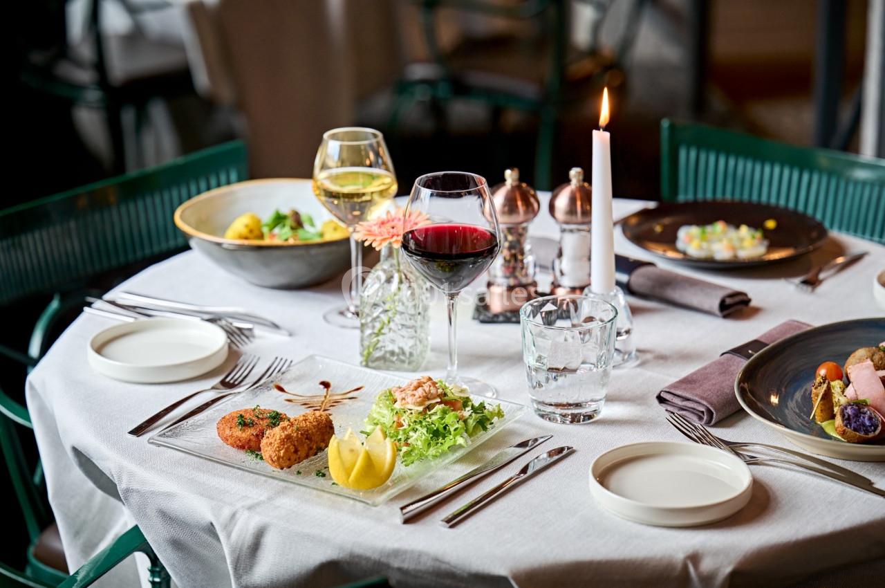 Table dressée avec des plats raffinés, verres de vin, bougie allumée et couverts sur une nappe blanche.