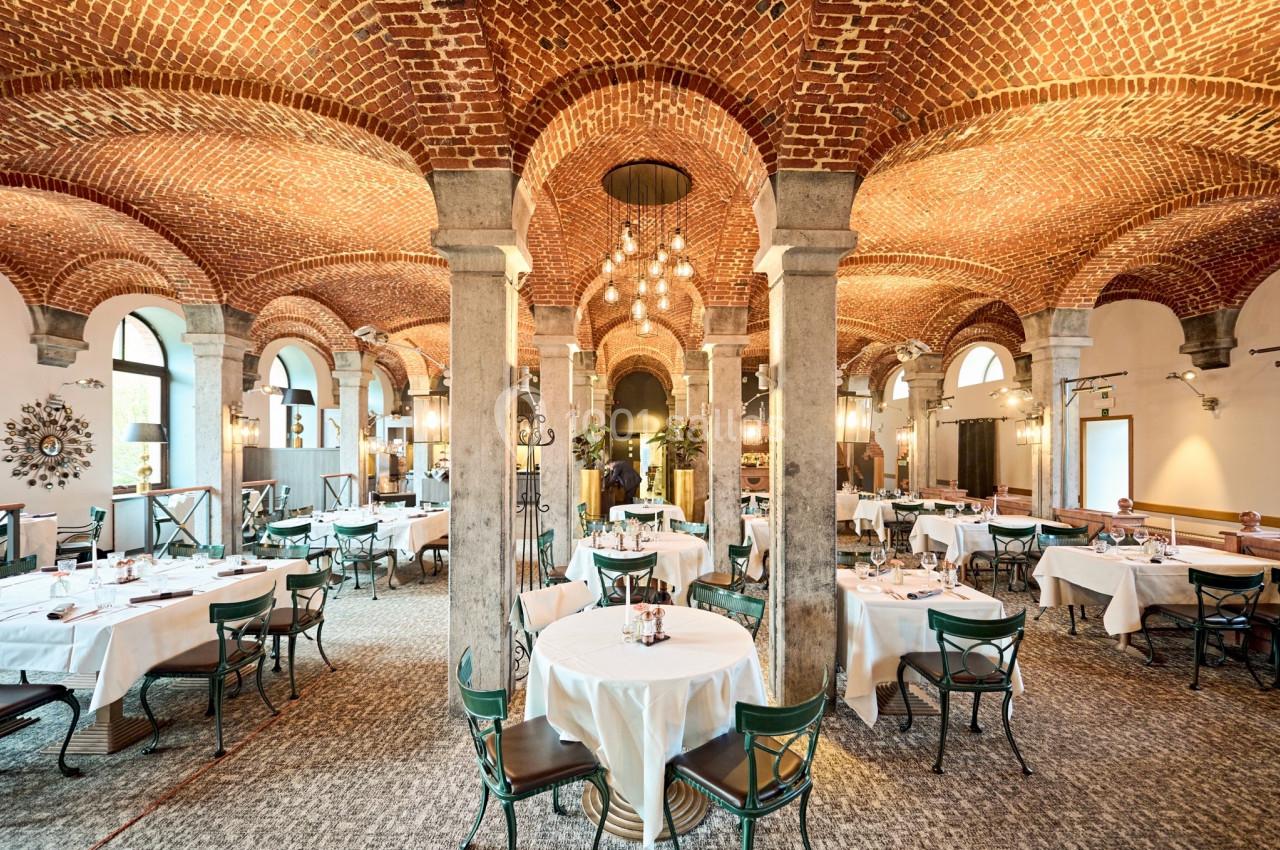 Salle de restaurant avec voûtes en briques, tables dressées avec nappes blanches et chaises en métal vert.