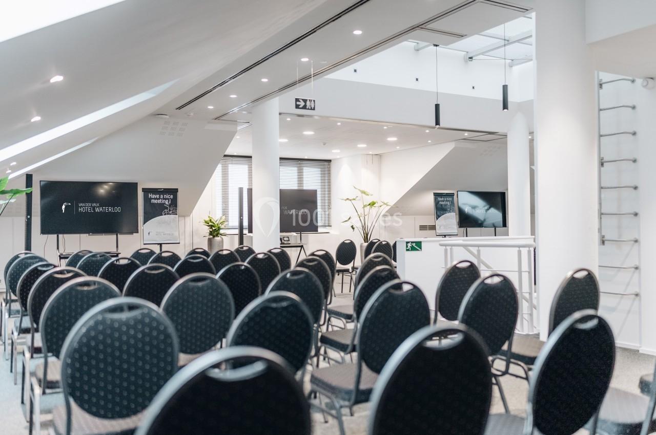 Salle de conférence moderne avec des rangées de chaises noires vides, écrans et éclairage naturel.
