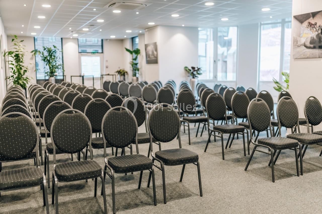 Salle de conférence lumineuse avec des rangées de chaises noires alignées et des plantes en arrière-plan.