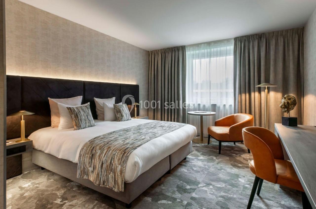 Chambre d'hôtel moderne avec lit double, fauteuils en cuir, bureau, éclairage doux et grande fenêtre avec rideaux.