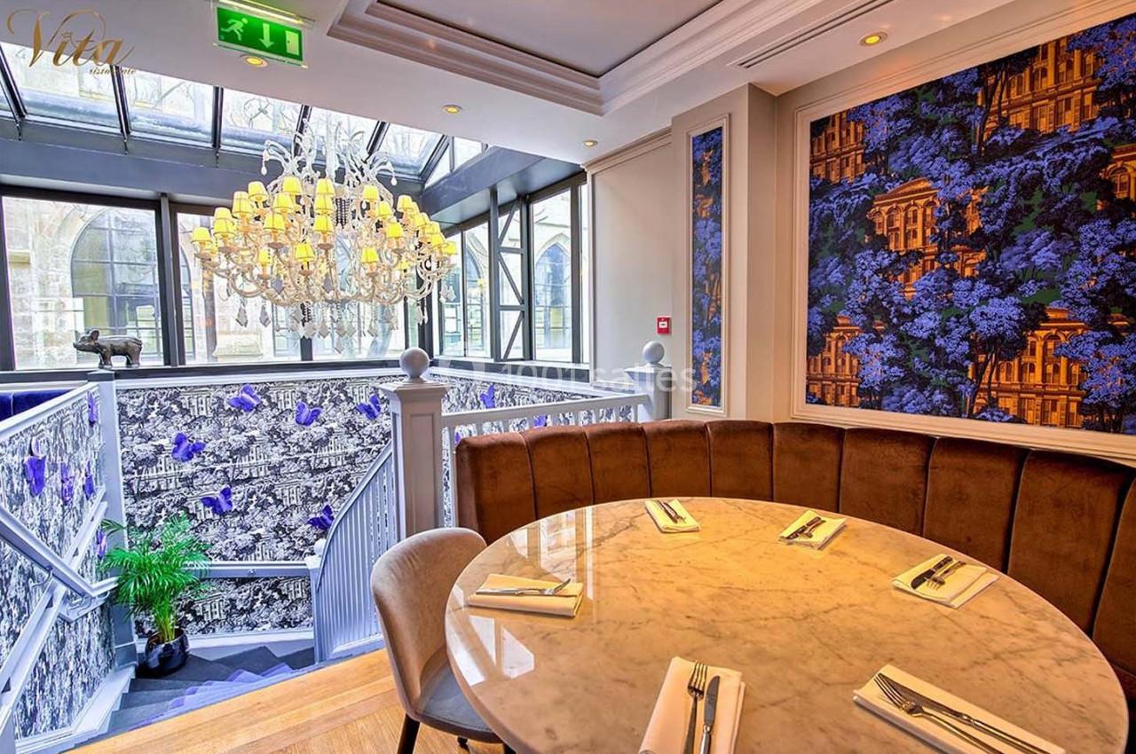 Salle de restaurant élégante avec table ronde, lustre imposant et décor floral sur les murs près d'un escalier.
