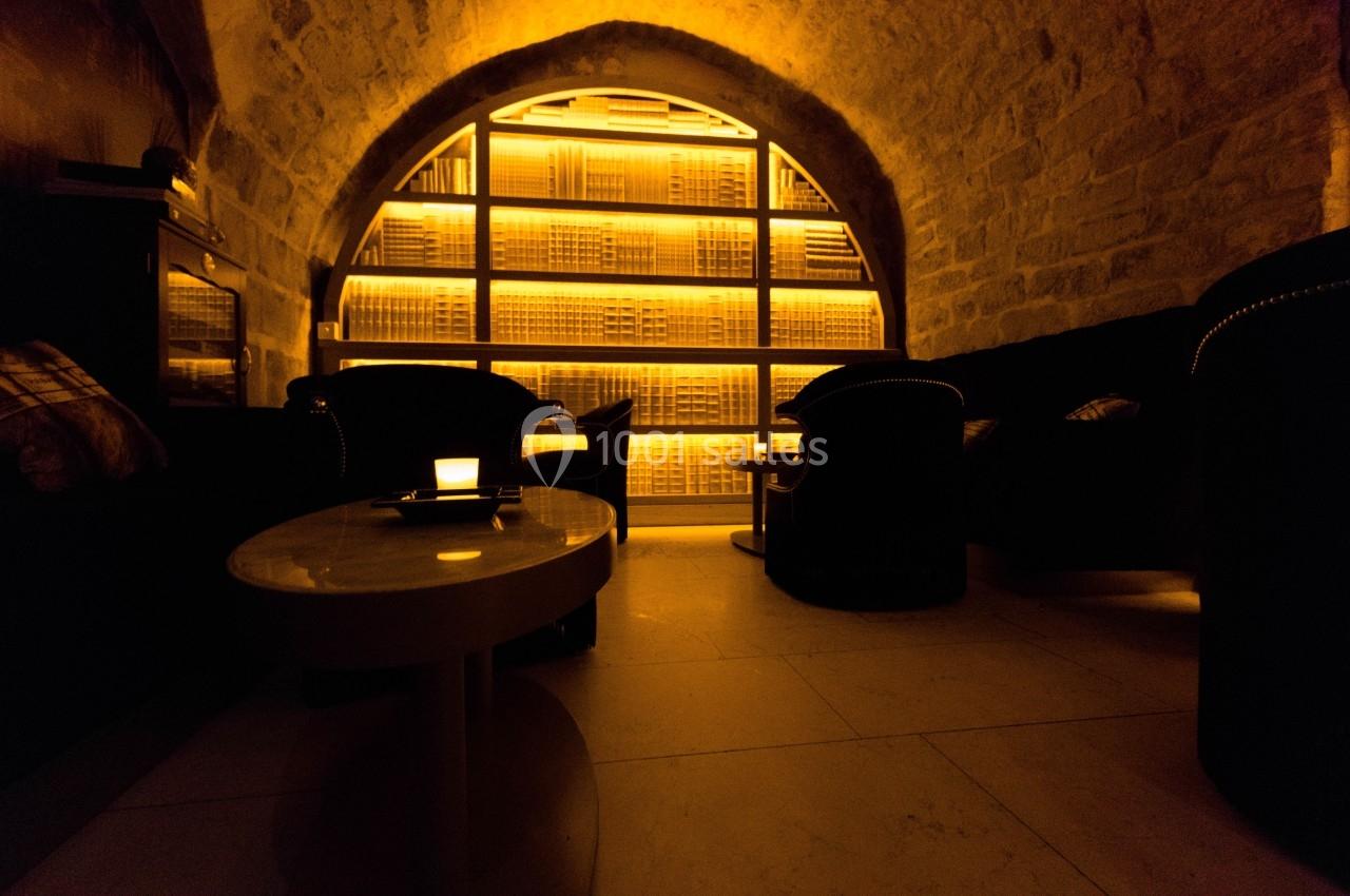 Salle voûtée avec éclairage tamisé, fauteuils sombres et étagères illuminées contenant des bouteilles.