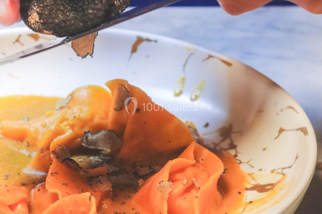 Des mains râpent une truffe noire sur des pâtes fraîches nappées de sauce dans une assiette blanche.