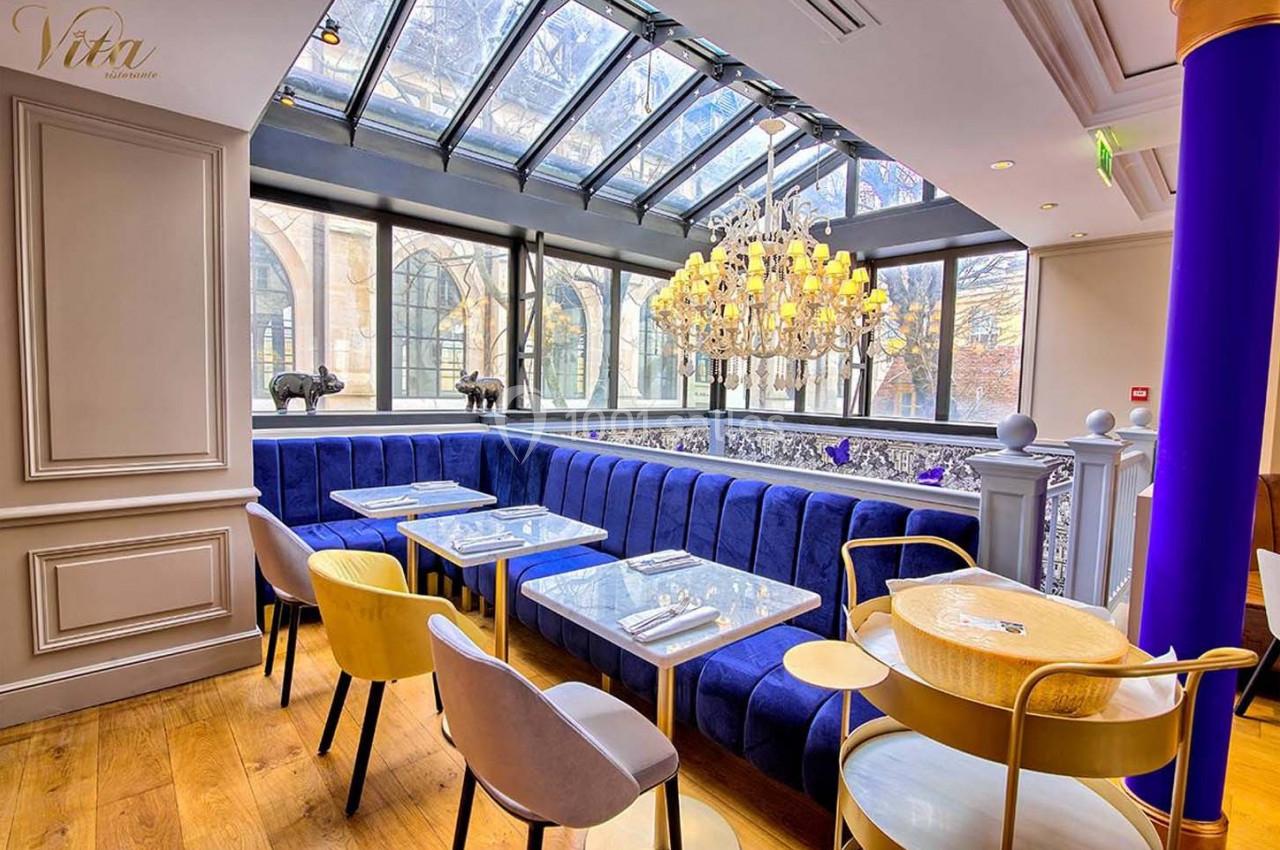 Salle de restaurant lumineuse avec verrière, banquettes bleues, chaises modernes et lustre élégant.