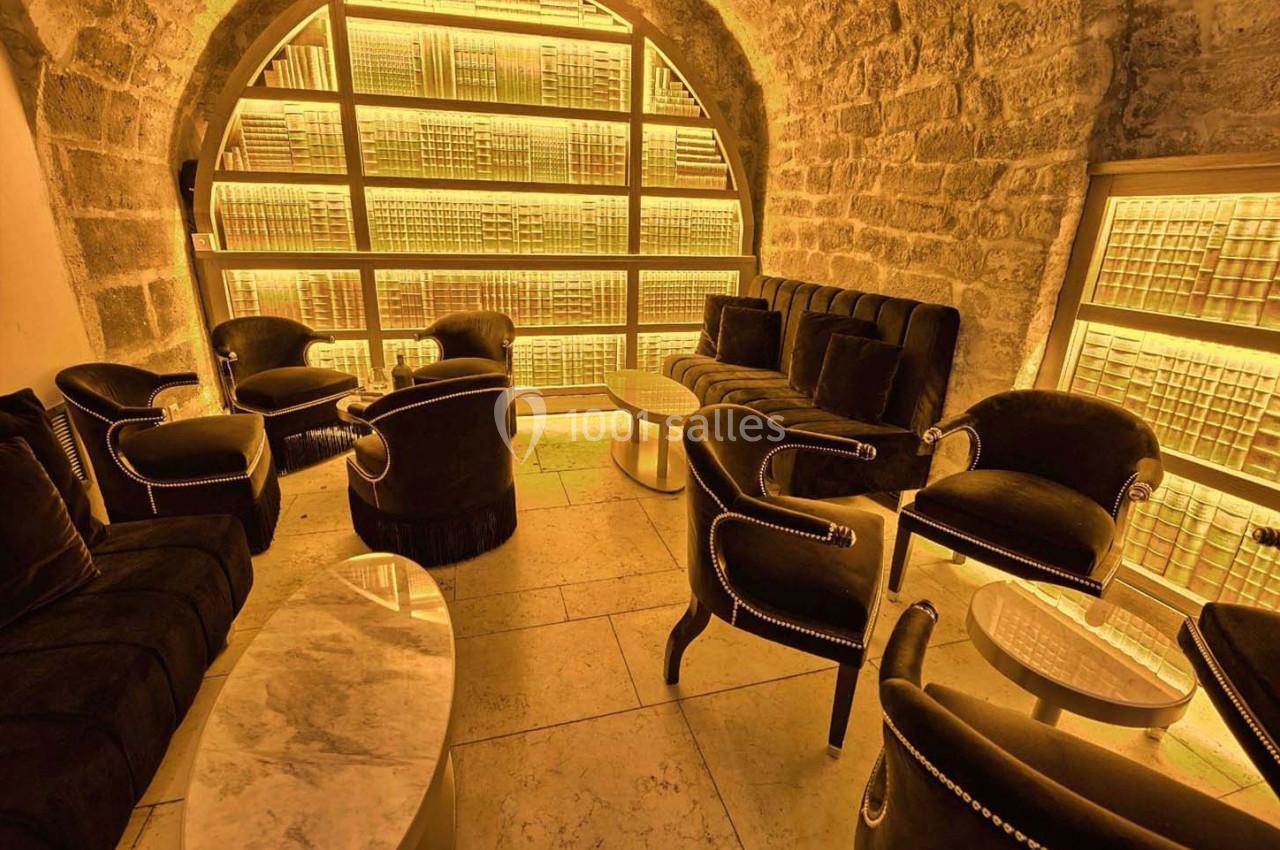 Salon voûté en pierre avec fauteuils noirs, tables basses en marbre et étagères illuminées en arrière-plan.