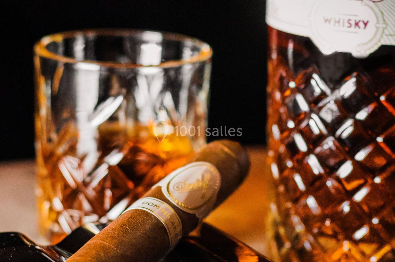Un cigare posé sur un cendrier noir, avec un verre de whisky et une bouteille en arrière-plan.