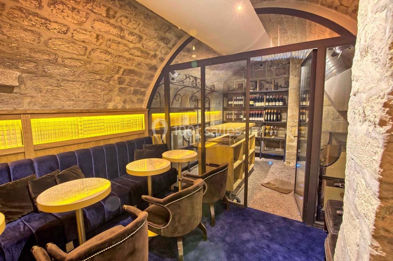 Salle voûtée en pierre avec tables, chaises en velours et étagères de bouteilles de vin derrière une cloison vitrée.