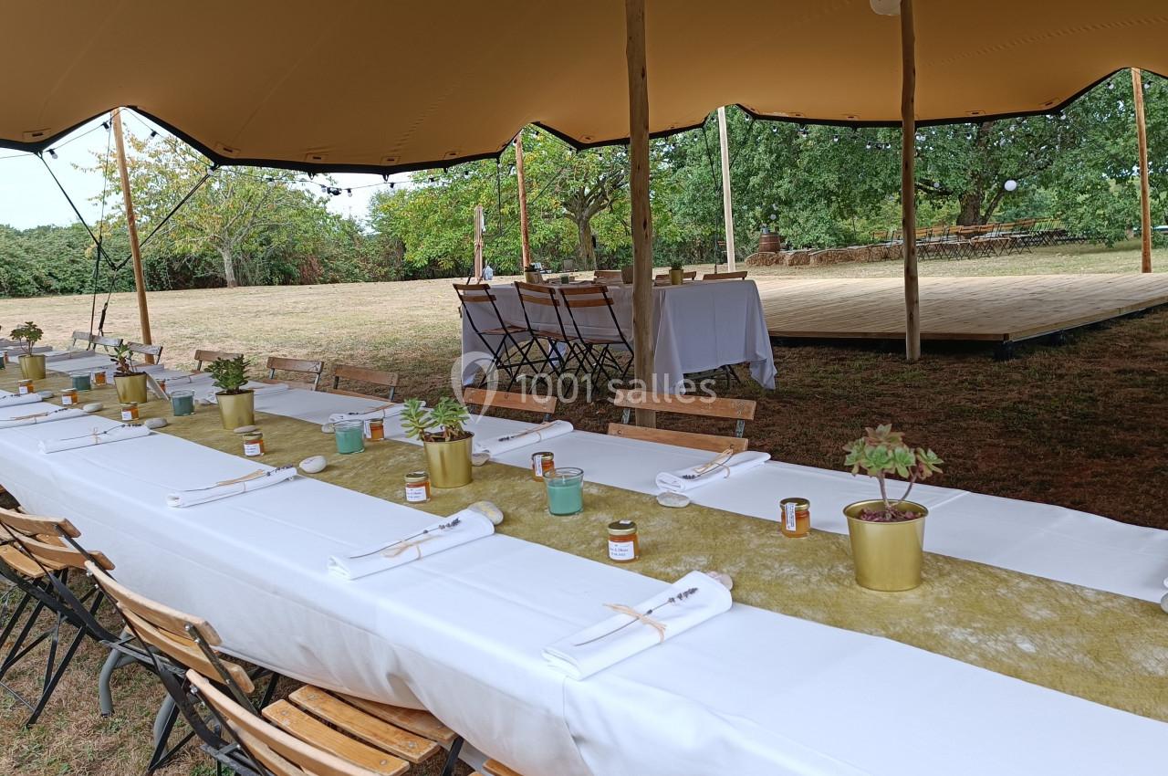 Tables dressées avec nappes blanches, plantes en pot et bougies, sous une tente dans un espace extérieur verdoyant.
