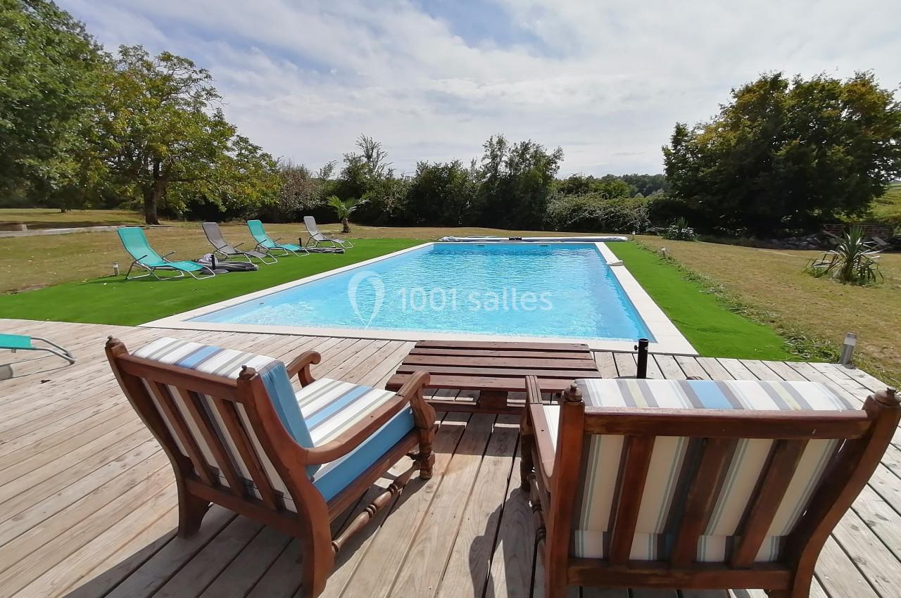 Deux fauteuils en bois sur une terrasse en bois font face à une piscine entourée de chaises longues et de verdure.