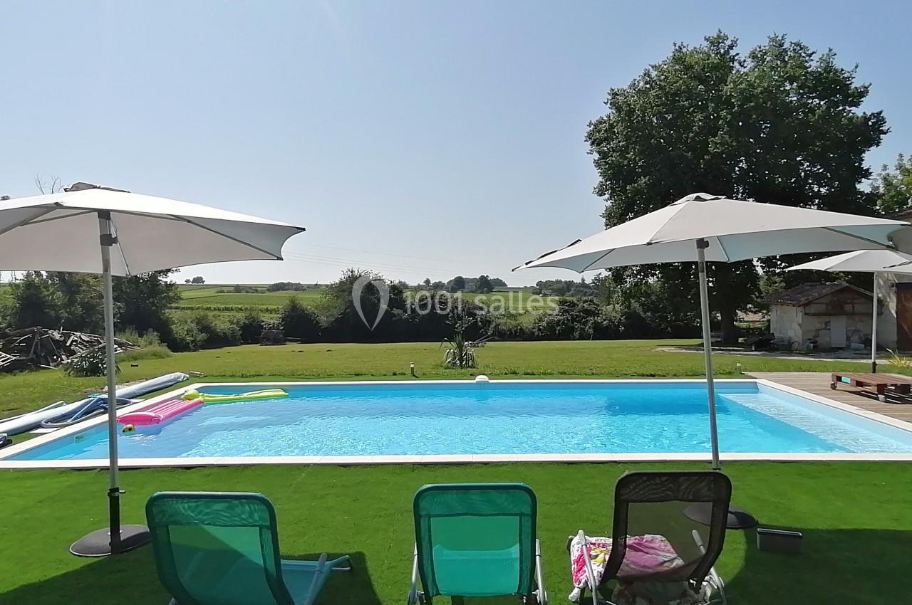 Piscine extérieure entourée de chaises longues, parasols et pelouse, avec vue sur la campagne en arrière-plan.