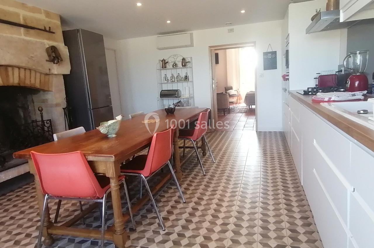 Cuisine lumineuse avec une grande table en bois, des chaises rouges, une cheminée en pierre et un sol à motifs géométriques.