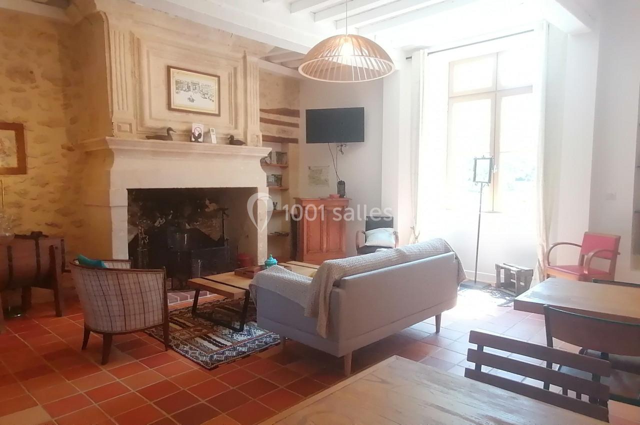 Salon lumineux avec cheminée en pierre, canapé, fauteuils, meubles en bois et grande fenêtre laissant entrer la lumière.
