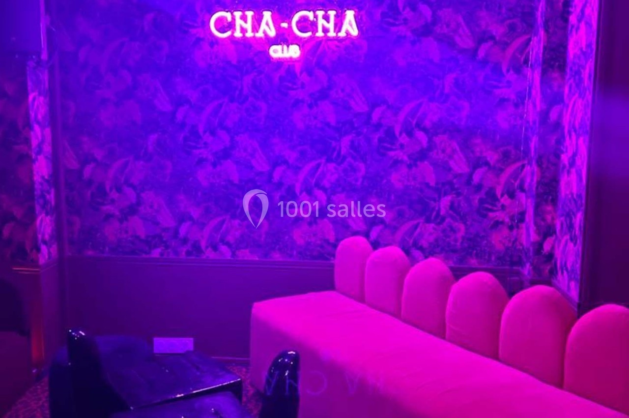 Banquette rose et fauteuil noir dans une pièce éclairée par une lumière violette avec un néon ’CHA-CHA Club’ au mur.