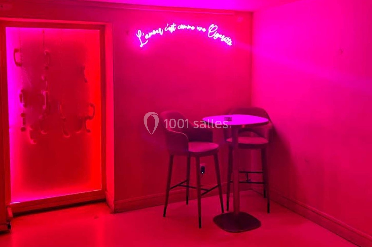 Deux chaises hautes et une table ronde dans une pièce éclairée par une lumière rose avec un néon au mur.
