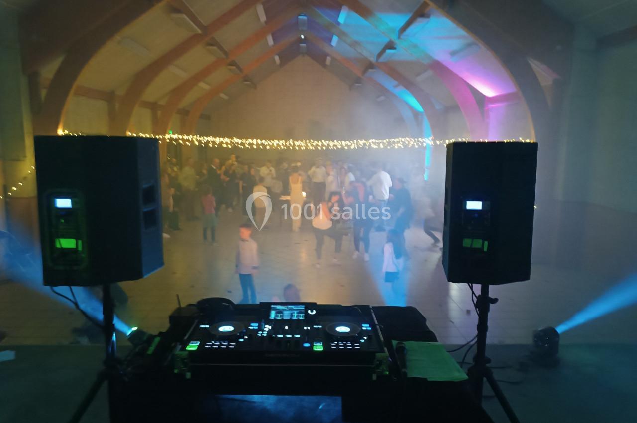 Vue depuis une table de mixage avec des enceintes, éclairage coloré et une foule dansante dans une salle festive.