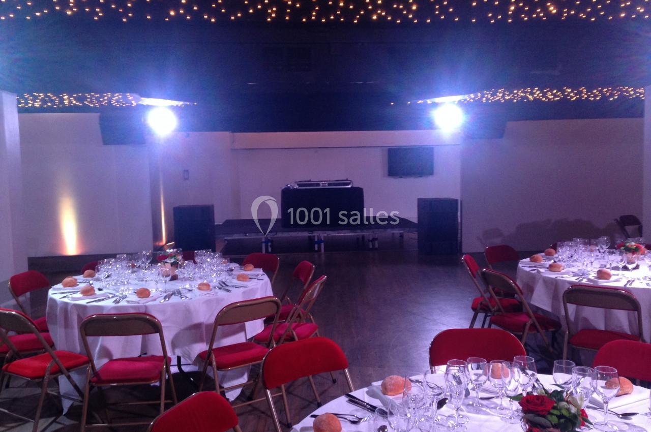 Salle de réception avec tables dressées, chaises rouges et une scène équipée d'un pupitre de DJ sous un plafond illuminé.