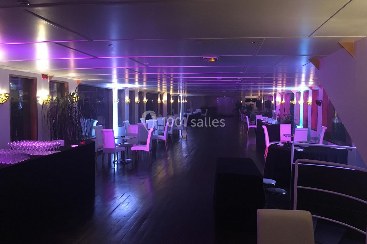 Salle de réception élégante avec tables et chaises blanches, éclairée par des lumières violettes.