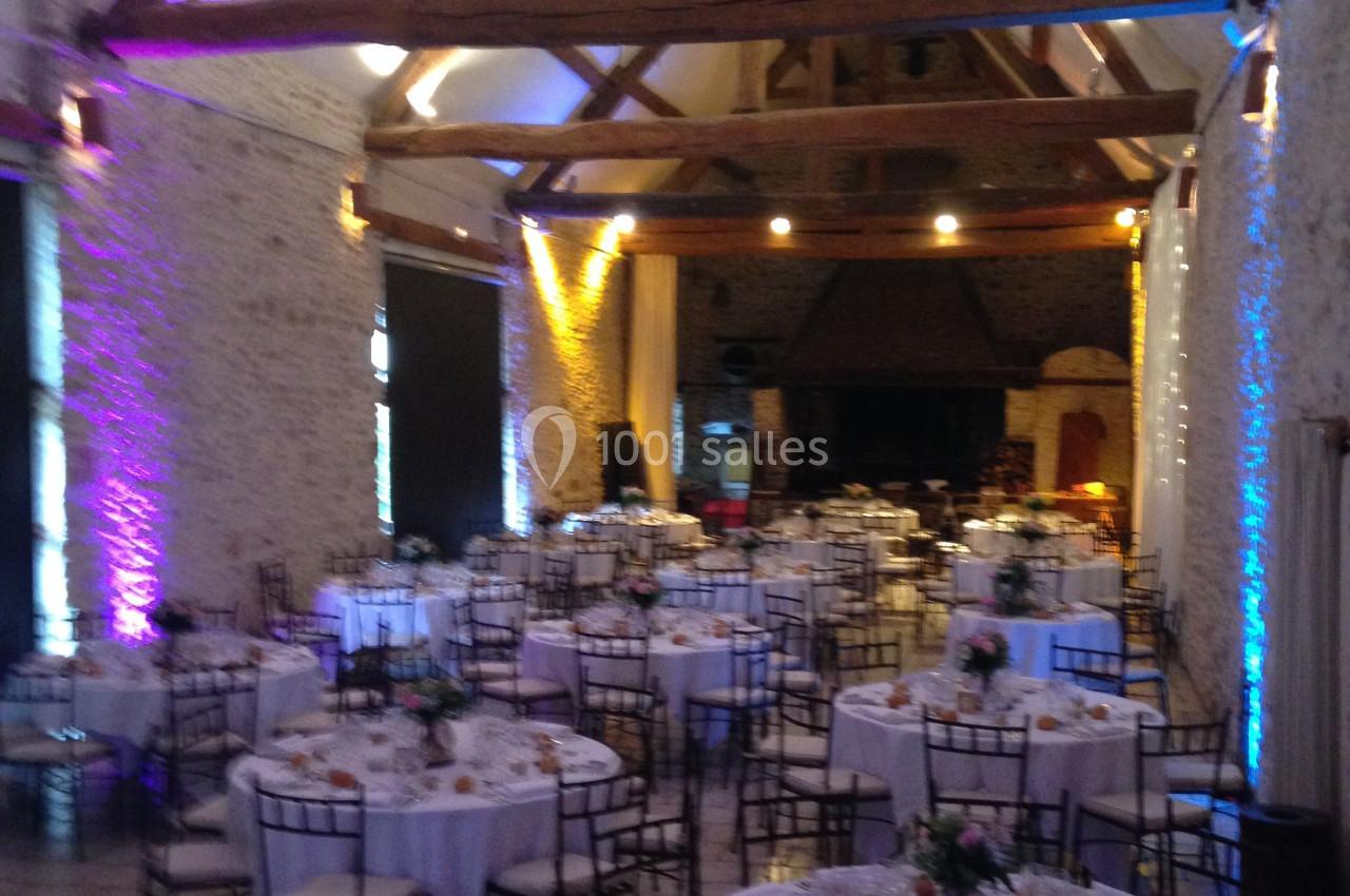 Salle de réception avec des tables rondes dressées, décorée de poutres apparentes et éclairages colorés.