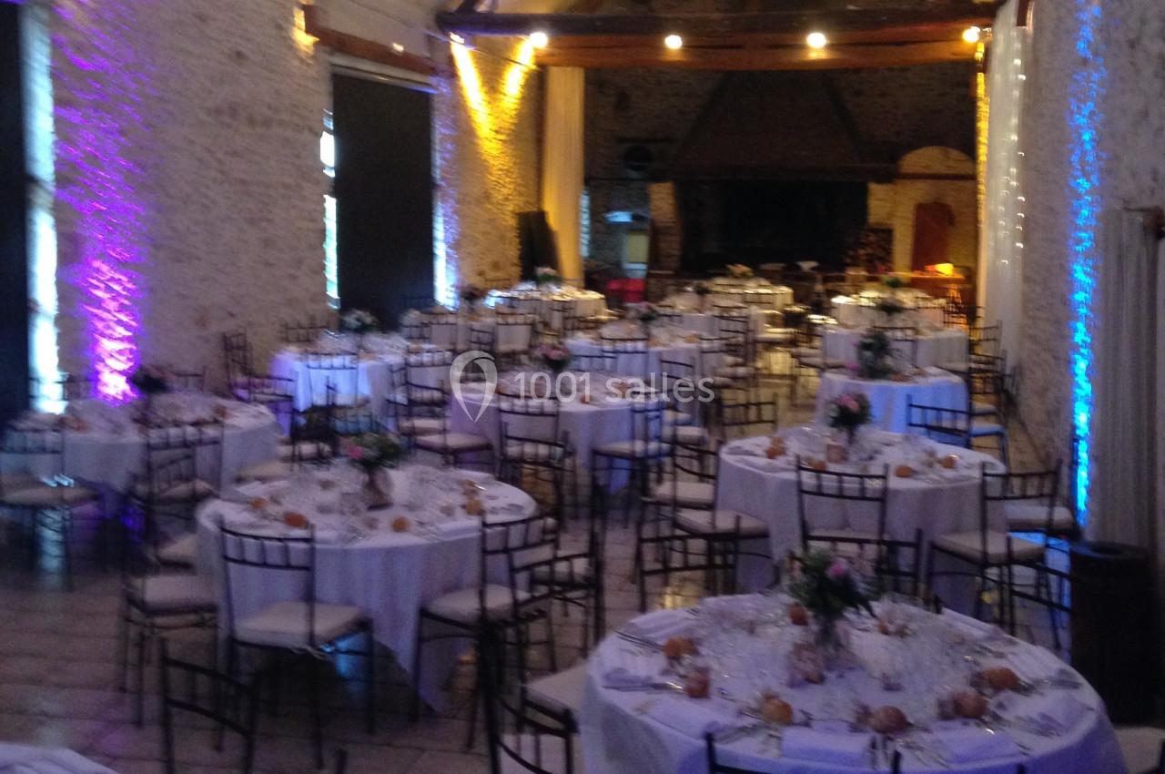 Salle de réception avec des tables rondes dressées, nappes blanches, chaises en métal et éclairage coloré.