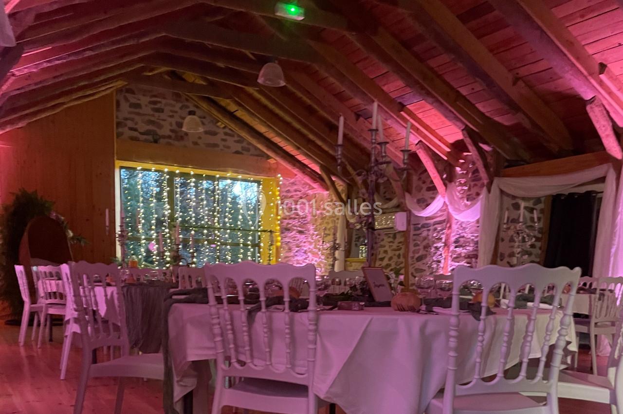 Salle de réception décorée avec des guirlandes lumineuses, des tables dressées et un plafond en poutres apparentes.