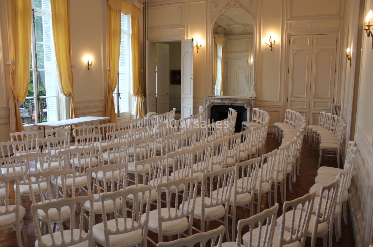 Salle lumineuse avec des rangées de chaises blanches alignées, murs ornés de moulures et grandes fenêtres avec rideaux…