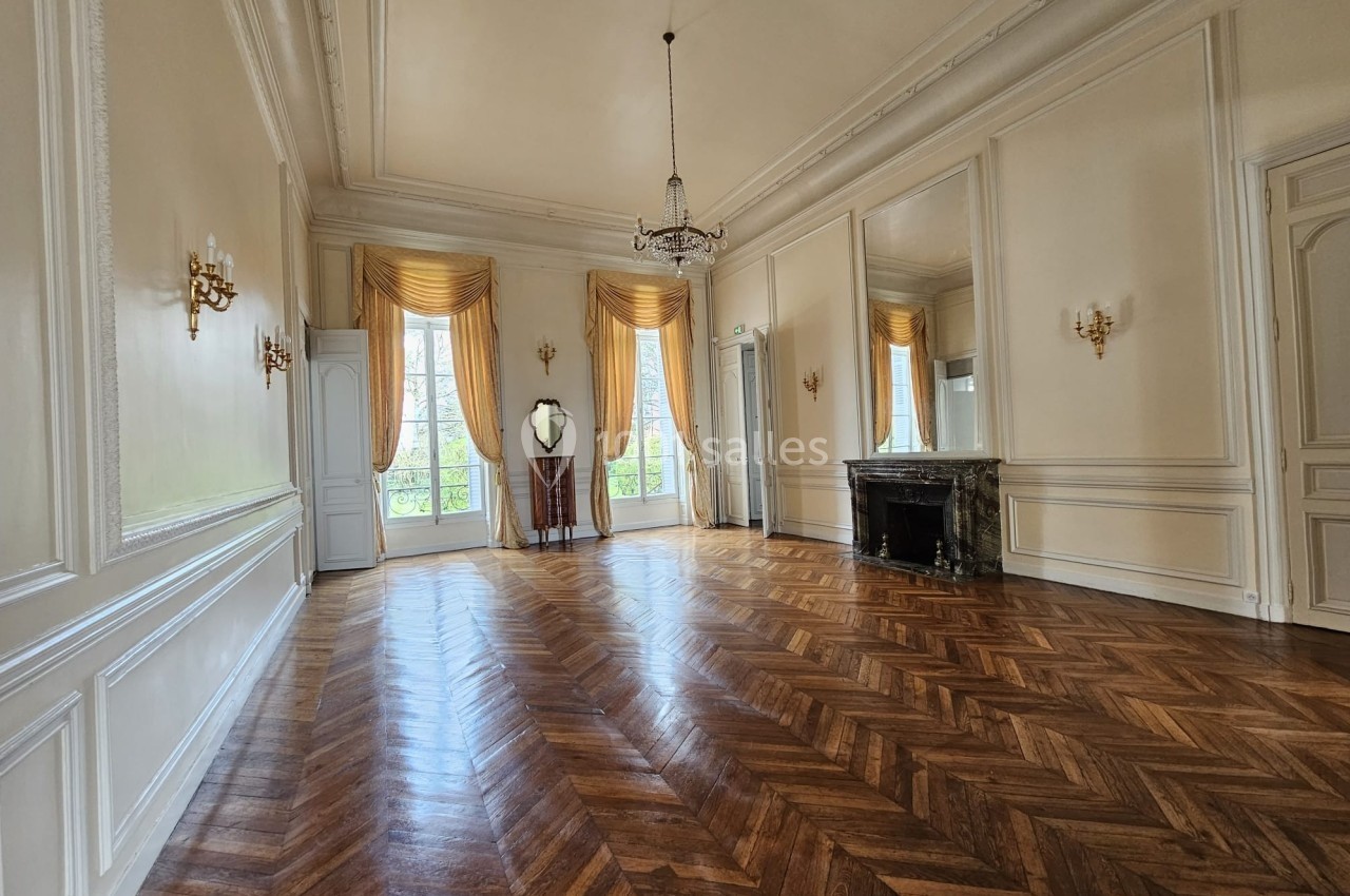 Salle lumineuse avec parquet en chevrons, grandes fenêtres, rideaux dorés, cheminée en marbre et miroirs muraux.