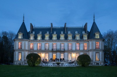 Façade d'un château illuminé la nuit avec une fontaine ornée de statues au premier plan.