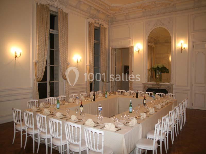 Salle élégante avec une grande table en U dressée, entourée de chaises blanches, éclairée par des appliques murales.
