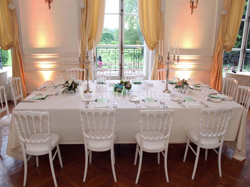 Table élégamment dressée avec nappes blanches, chandeliers, vaisselle raffinée et chaises blanches dans une salle lumineuse.