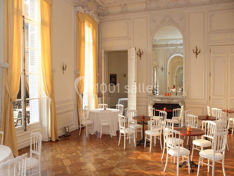 Salle lumineuse avec parquet, chaises blanches, tables rondes et grandes fenêtres encadrées de rideaux jaunes.