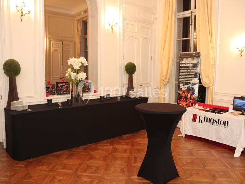 Salle élégante avec parquet, table drapée noire ornée de fleurs blanches et stand promotionnel en arrière-plan.