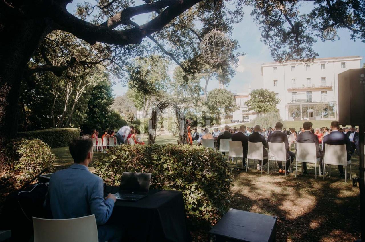 Cérémonie en plein air sous des arbres, avec des invités assis et une personne à une table équipée d'un ordinateur.