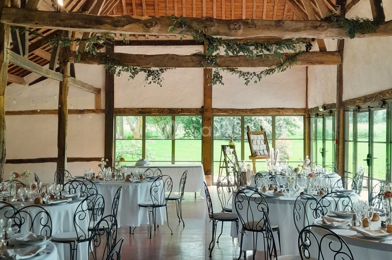 Salle de réception lumineuse avec tables rondes dressées, chaises en fer forgé et poutres apparentes en bois.