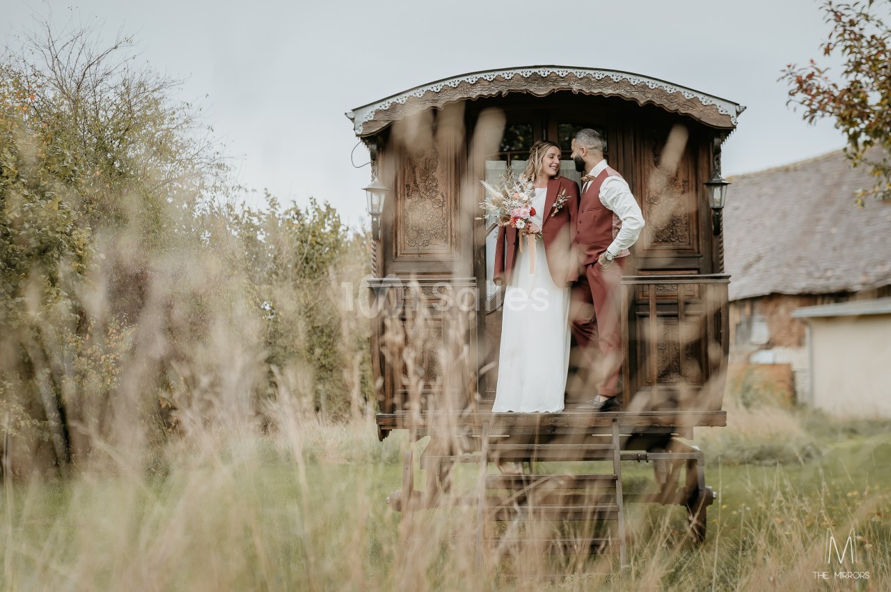 Un couple en tenue de mariage pose sur la plateforme d'une roulotte en bois dans un cadre champêtre.