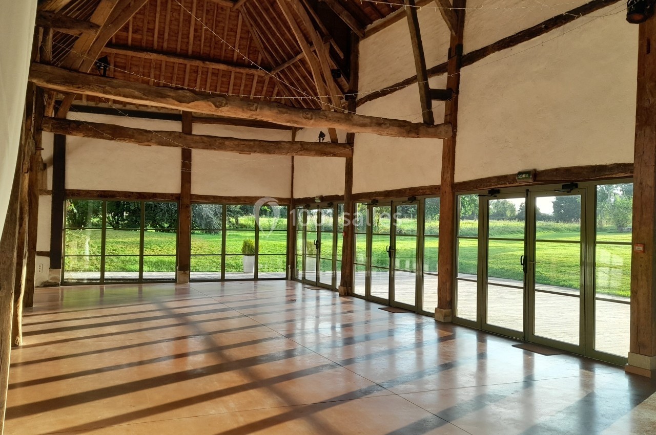 Intérieur d'une grande salle avec charpente en bois, baies vitrées et vue sur un espace vert ensoleillé.
