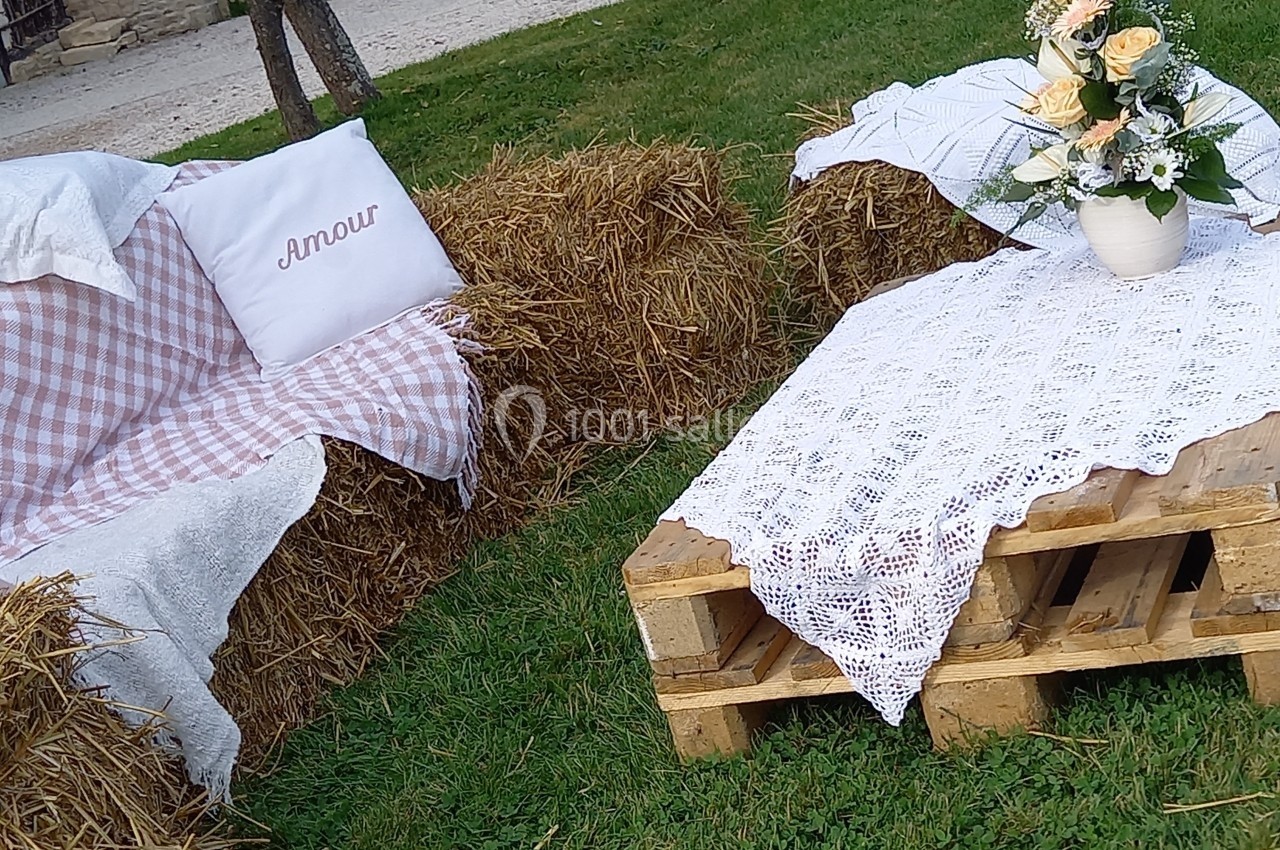 Canapé en bottes de paille avec coussin et nappe à carreaux, table en palettes décorée de dentelle et d'un bouquet de fleurs.
