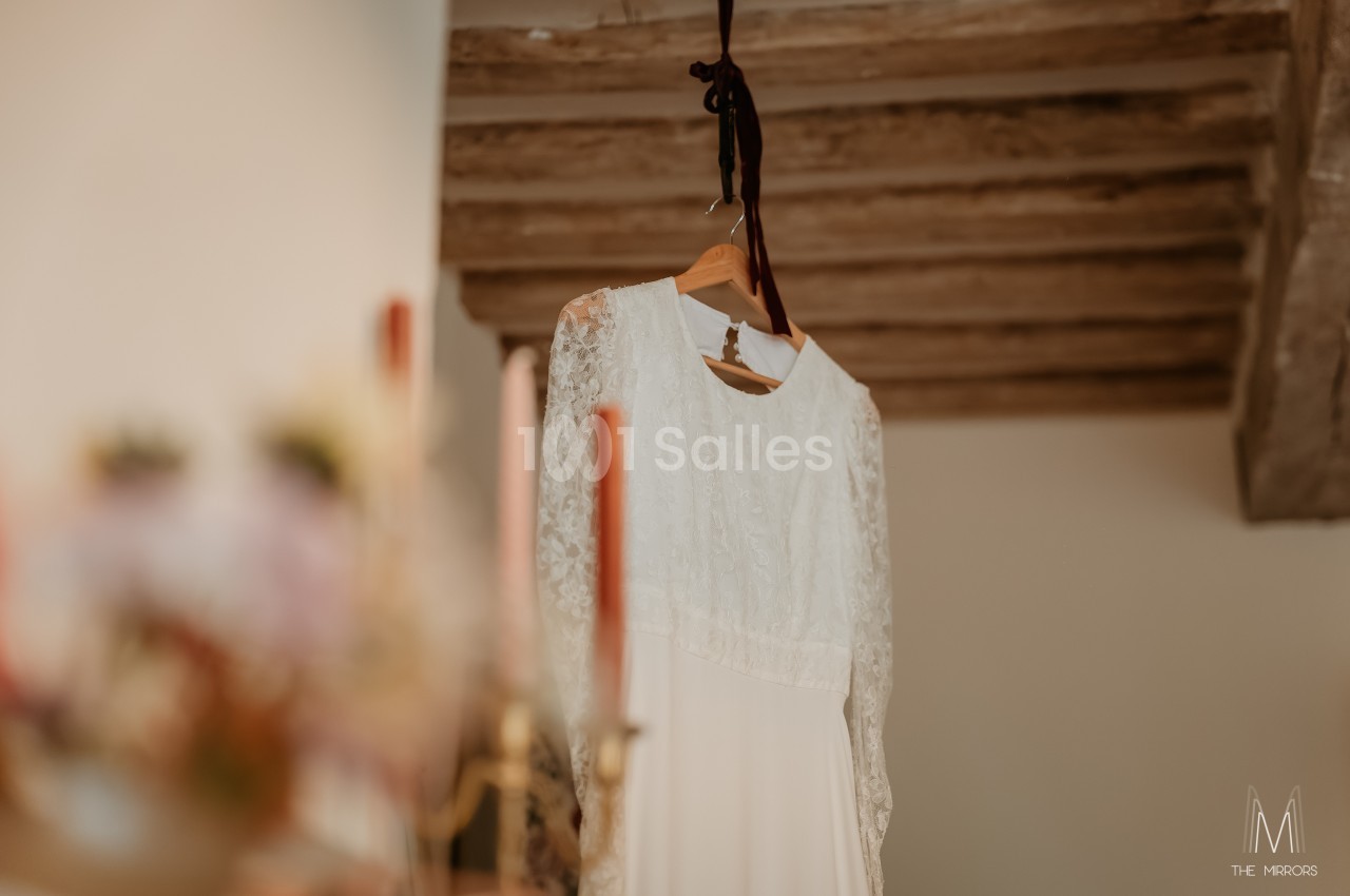 Robe de mariée blanche en dentelle suspendue sur un cintre, avec des poutres en bois en arrière-plan.