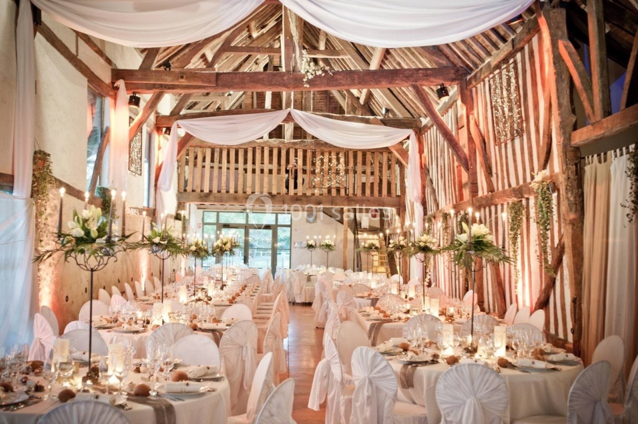 Salle de réception décorée avec des tables dressées, des chaises blanches et des drapés suspendus au plafond en bois.