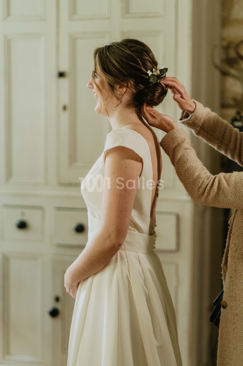 Une femme en robe de mariée blanche se fait ajuster la coiffure par une autre personne dans une pièce lumineuse.