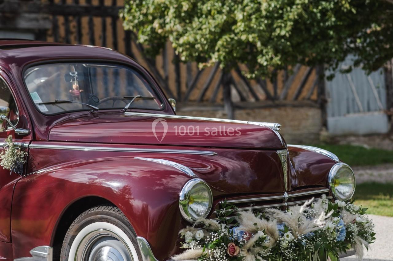 Voiture ancienne rouge décorée de fleurs blanches et feuillages, stationnée devant un bâtiment à colombages.